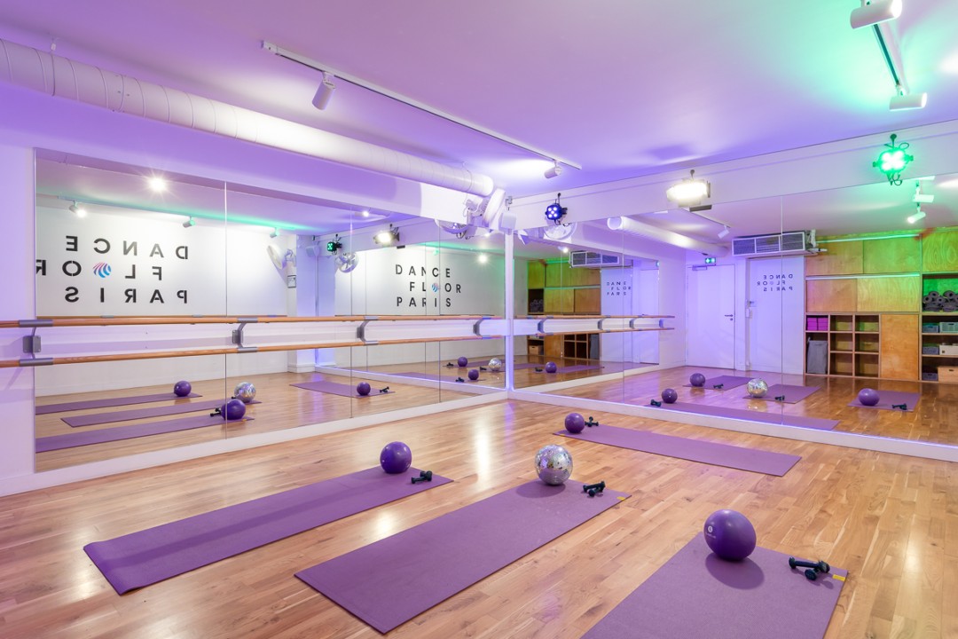 Salle de danse Dancefloor Paris au parquet clair, avec tapis violets, petits ballons, miroirs et barres, logo Dancefloor Paris au mur