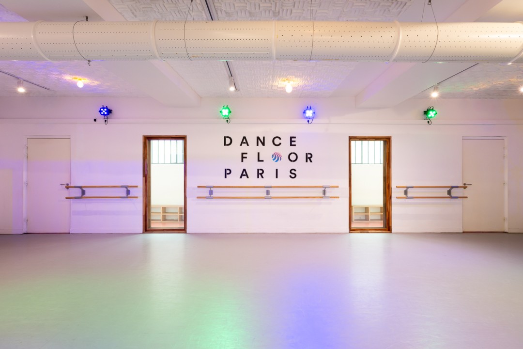 Salle de danse vide aux murs blancs, barres en bois et projecteurs bleus et verts, logo Dancefloor Paris au mur